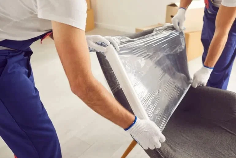 Two professional movers in white gloves carefully wrapping upholstered furniture in protective plastic during a white glove moving service